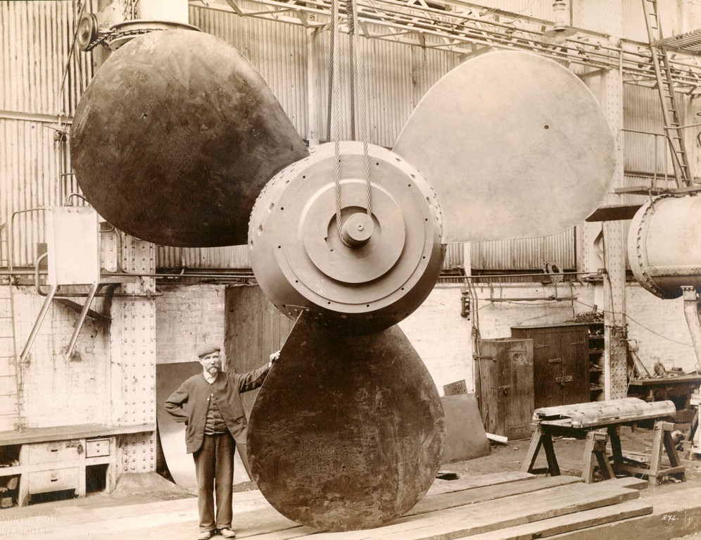  Black and white photograph of a man standing next to a large propeller